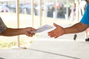 One person passes a fundraising catalog to another outdoors, both reaching out as they share opportunities to support their cause.