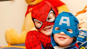 Two children in Spider-Man and Captain America costumes smile indoors, sitting together with a large teddy bear behind them—perfect for fundraising flyers.