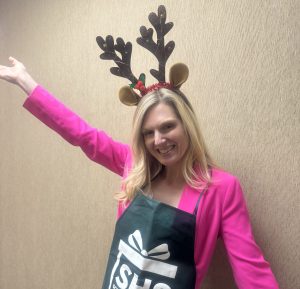 A cheerful individual in reindeer antlers, pink blazer, and green apron celebrates our fundraising spirit against a beige wall.