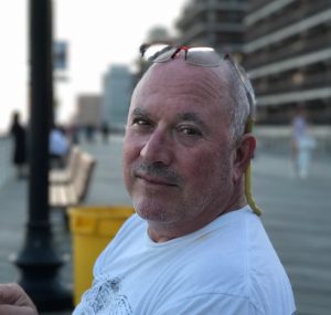 A friendly middle-aged man with glasses sits on a boardwalk bench, inviting your school to join our fundraising program.